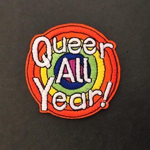 Peut inclure: Écusson brodé circulaire avec le texte "Queer All Year!" en blanc, bordé de noir, sur un fond arc-en-ciel. Le patch a une bordure extérieure orange.