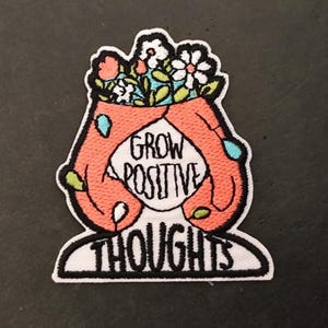 Peut inclure: Écusson brodé avec une bordure blanche. Il présente une tête stylisée avec des cheveux et des mains corail, et les mots "GROW POSITIVE THOUGHTS" dans un cercle blanc. La tête est surmontée d'un bouquet de fleurs.
