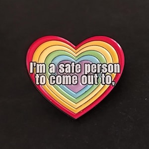 Op de afbeelding: Een hartvormige pin met een regenboogontwerp en de tekst "I'm a safe person to come out to." De pin heeft een rode rand en concentrische lagen regenboogkleuren, waaronder rood, oranje, geel, groen, blauw en paars.