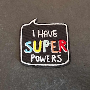 Peut inclure: Patch brodé noir et blanc avec une bulle de dialogue. Le texte à l'intérieur de la bulle de dialogue est "I have super powers" avec le mot "super" en couleurs arc-en-ciel.