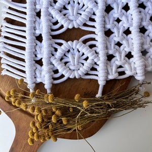 Può includere: Arazzo da parete in macramè bianco con un motivo geometrico, con un bordo a frangia. L'arazzo è esposto su una superficie in legno con fiori gialli secchi.