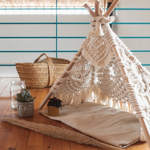 Op de afbeelding: Een witte macramé tipi tent voor huisdieren, met een houten frame en een wit katoenen kussen erin. De tipi staat op een houten vloer met een rieten mand en potplanten.