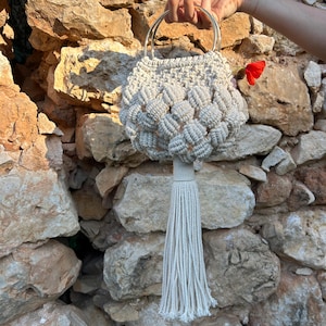 May include: A cream-colored macrame handbag with a round shape and a long tassel. The bag has two silver metal handles and a woven pattern. A red flower is tucked near the handle. The bag is held in front of a stone wall.
