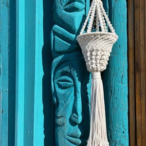 May include: White macrame plant hanger with a tassel hanging from a wooden tiki statue against a teal blue wall.