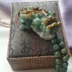 May include: A jade carving of a mythical creature in shades of green, brown, and white, resting on a decorative box. A strand of green jade beads hangs down. The box has a dark patterned top and a brown woven border.