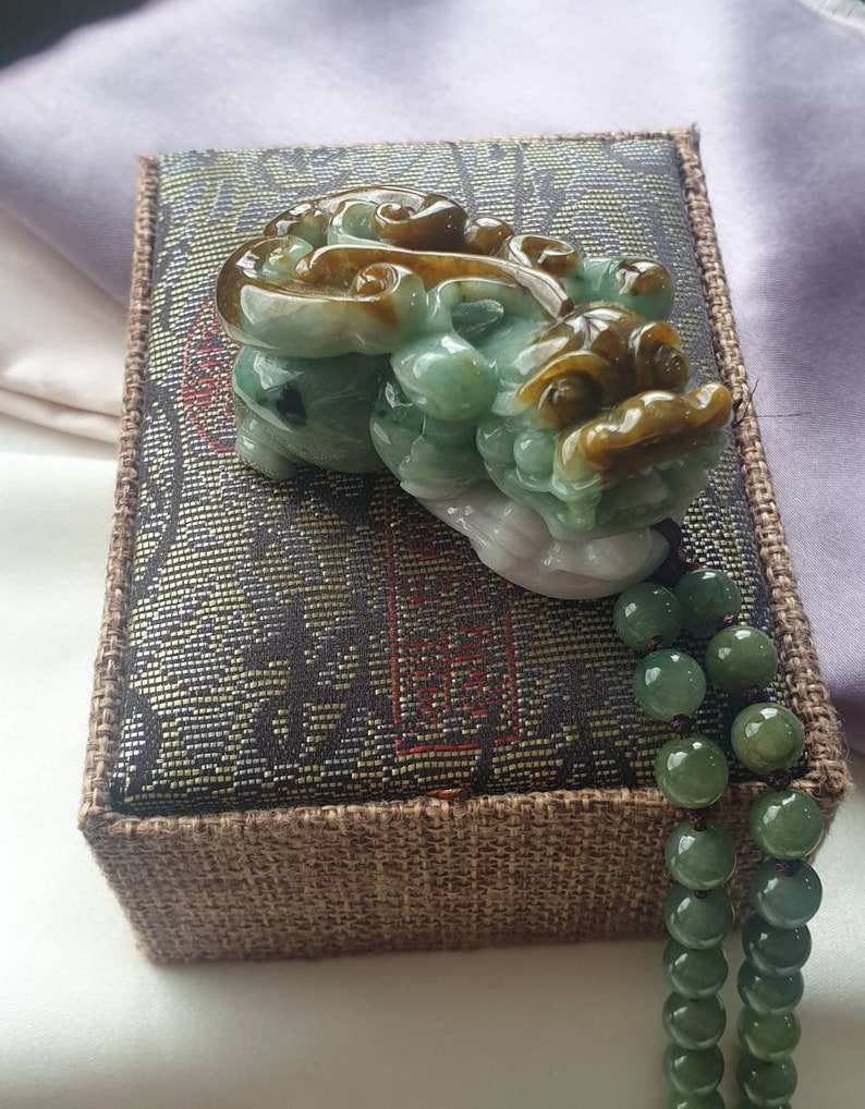 May include: A jade carving of a mythical creature in shades of green, brown, and white, resting on a decorative box. A strand of green jade beads hangs down. The box has a dark patterned top and a brown woven border.
