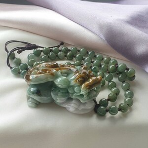 May include: A jade necklace featuring a carved pendant of a mythical creature in shades of green and brown. The pendant is attached to a beaded necklace with green beads. The necklace rests on a white and lavender satin surface.