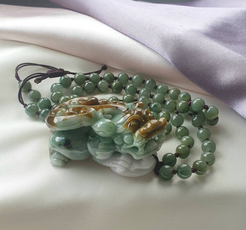 May include: A jade necklace featuring a carved pendant of a mythical creature in shades of green and brown. The pendant is attached to a beaded necklace with green beads. The necklace rests on a white and lavender satin surface.