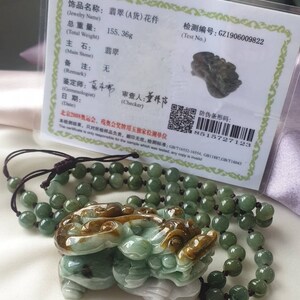May include: A jade pendant carved in the shape of a mythical creature, with shades of green, brown, and white. It is strung on a beaded necklace. A certificate with Chinese text is in the background, with a total weight of 155.36 grams.
