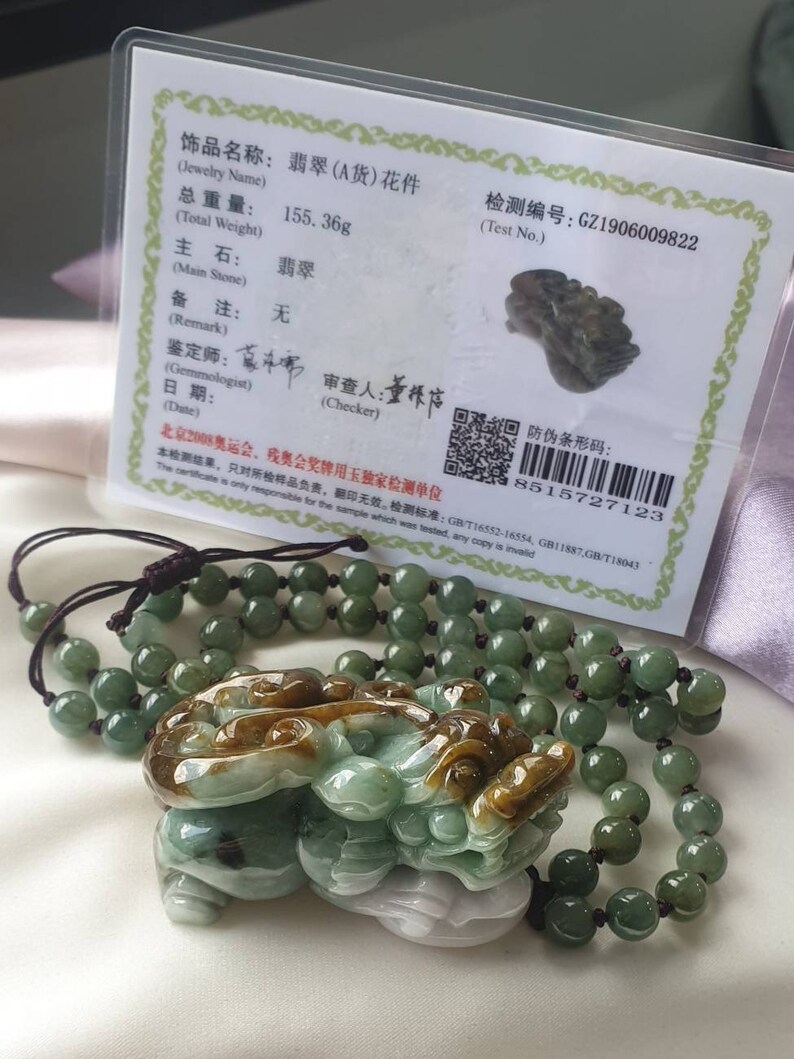 May include: A jade pendant carved in the shape of a mythical creature, with shades of green, brown, and white. It is strung on a beaded necklace. A certificate with Chinese text is in the background, with a total weight of 155.36 grams.