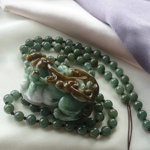 May include: A jade necklace featuring a carved green and brown mythical creature pendant. The pendant is surrounded by a beaded necklace of green jade spheres. The necklace is displayed on a white and purple fabric.