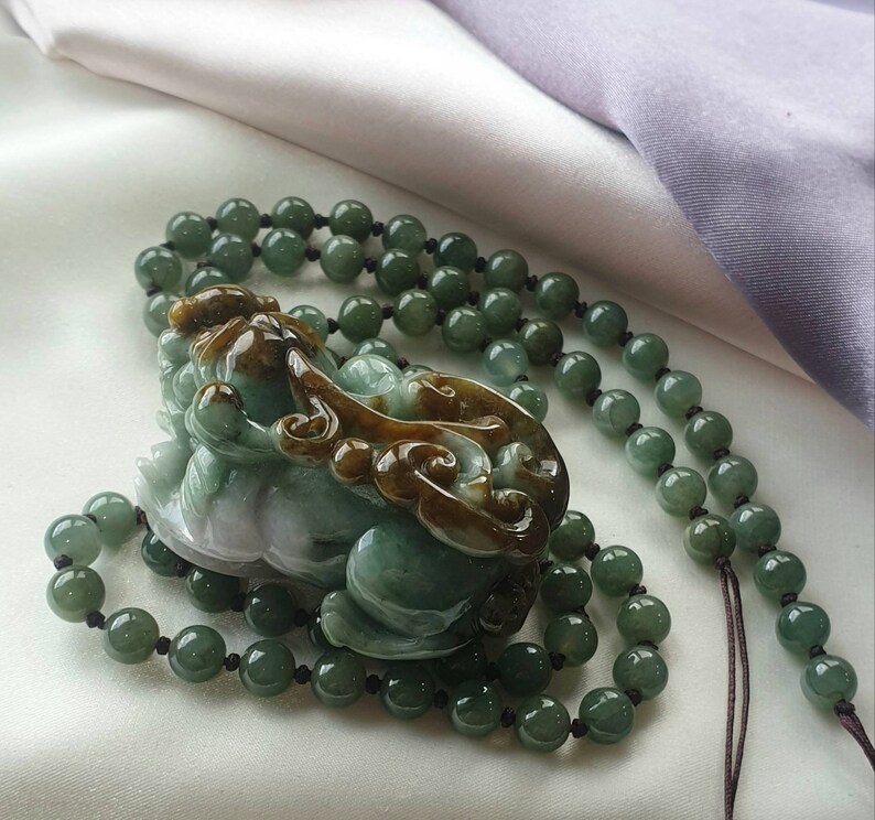 May include: A jade necklace featuring a carved green and brown mythical creature pendant. The pendant is surrounded by a beaded necklace of green jade spheres. The necklace is displayed on a white and purple fabric.