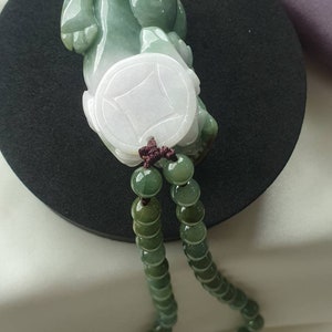 May include: A jade pendant featuring a carved figure with a coin design, in shades of green and white. The pendant is strung on a beaded necklace with a dark brown cord. The jade has a polished finish, and the beads are a gradient of green.