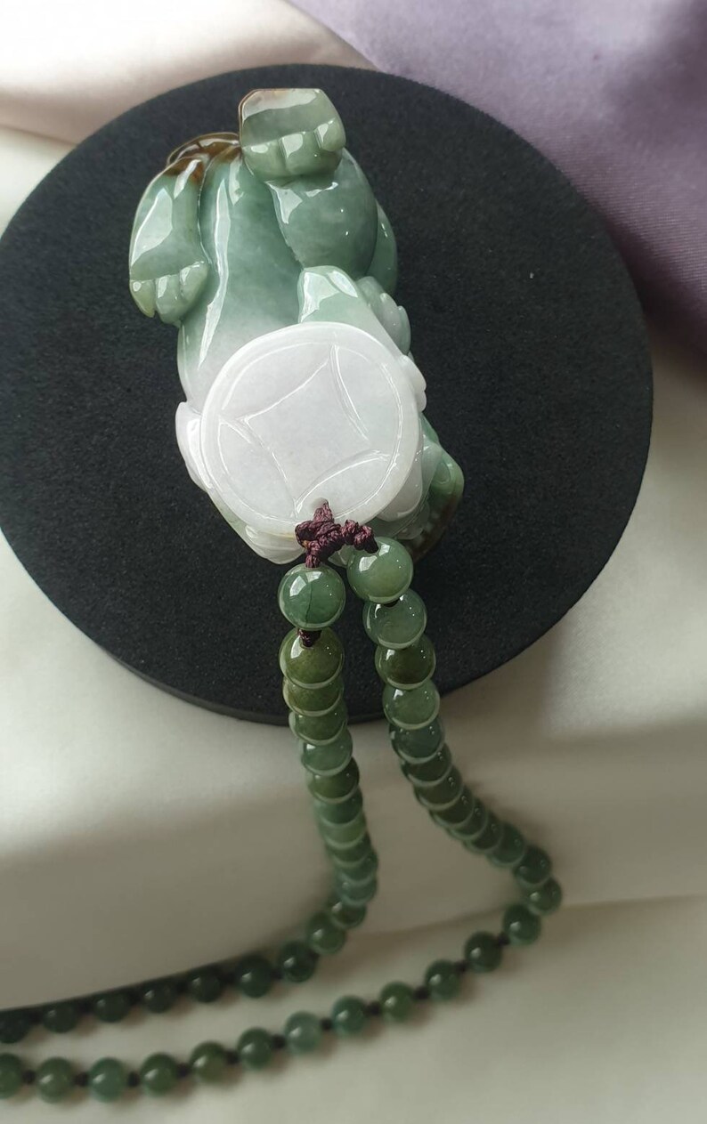 May include: A jade pendant featuring a carved figure with a coin design, in shades of green and white. The pendant is strung on a beaded necklace with a dark brown cord. The jade has a polished finish, and the beads are a gradient of green.