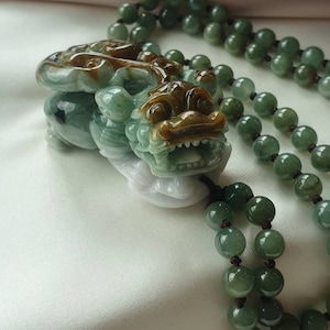 May include: A jade pendant necklace featuring a detailed carving of a mythical creature with a fierce expression. The pendant is green and brown, with a matching beaded necklace. The beads are a deep green color.