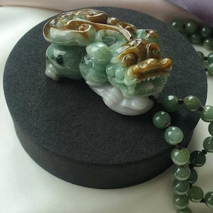 May include: A jade carving of a mythical creature in shades of green, brown, and white, resting on a black surface. The detailed sculpture features a dragon-like form with intricate carvings. A strand of green jade beads is visible in the background.