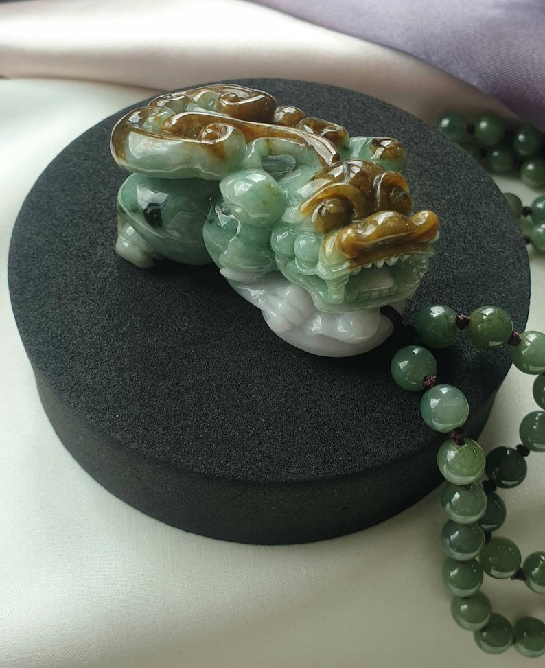 May include: A jade carving of a mythical creature in shades of green, brown, and white, resting on a black surface. The detailed sculpture features a dragon-like form with intricate carvings. A strand of green jade beads is visible in the background.