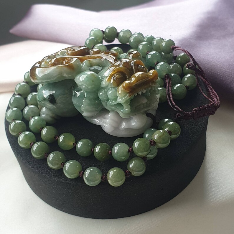 May include: A jade pendant featuring a detailed carving of a mythical creature, in shades of green, brown, and white. The pendant is accompanied by a beaded necklace with green beads. The piece is displayed on a black stand.