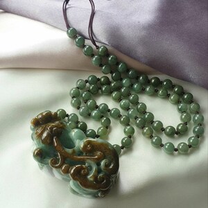 May include: A jade necklace with a carved pendant. The necklace features round, green beads strung on a dark brown cord. The pendant is a detailed carving in shades of green and brown.