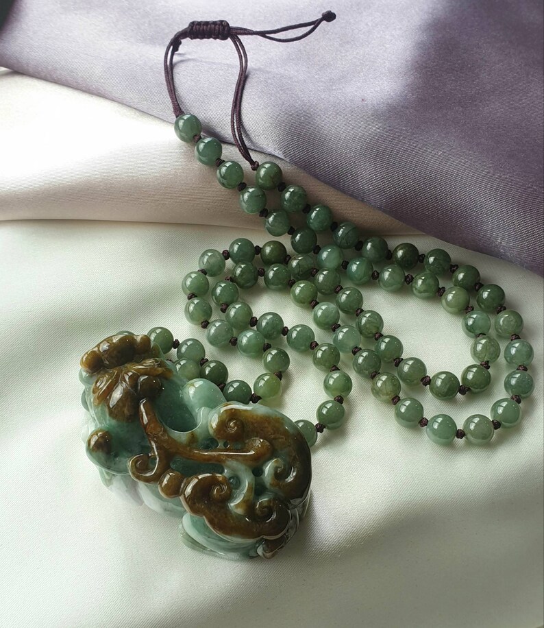 May include: A jade necklace with a carved pendant. The necklace features round, green beads strung on a dark brown cord. The pendant is a detailed carving in shades of green and brown.