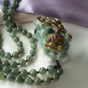 May include: A jade necklace with a carved pendant. The necklace features round, green beads strung on a dark cord. The pendant is a detailed carving of a mythical creature in shades of green and brown.