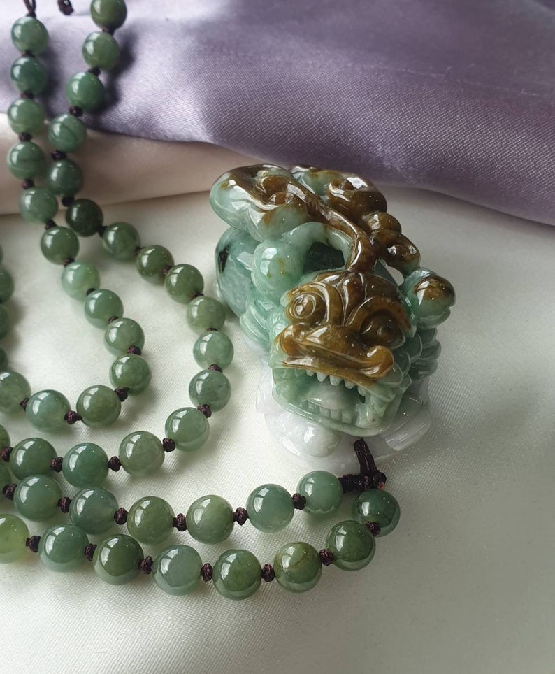 May include: A jade necklace with a carved pendant. The necklace features round, green beads strung on a dark cord. The pendant is a detailed carving of a mythical creature in shades of green and brown.