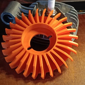 Könnte beinhalten: Hellorange Lüfterrad mit kreisförmigem Design und mehreren Lamellen. Das Lüfterrad befindet sich vor einer grauen Motorkomponente und einer Zündkerze. Das Lüfterrad ist wahrscheinlich ein Ersatzteil für einen kleinen Motor.