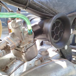 Könnte beinhalten: Nahaufnahme eines Motorradmotors mit Vergaser und einem schwarzen Lufteinlass mit sechseckigem Gitter. Der Motor ist aus Metall und hat eine silberne Oberfläche.