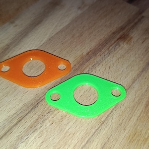 Könnte beinhalten: Zwei kleine, rautenförmige Objekte mit einem zentralen Loch und zwei kleineren Löchern. Eines ist leuchtend orange, das andere leuchtend grün. Sie liegen auf einer Holzoberfläche.