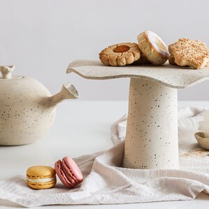 Still life in high-key. On the right is a beige marbled stoneware cake stand with several cookies on top. On the table, there is a beige cloth with several sweets on top. On the left is a teapot of the same material.