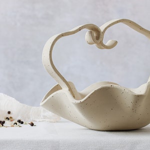 Front view of a mottled beige ceramic basket on a table with a white tablecloth. It has a spiral handle in the center. A glass salt grinder can be seen behind it.