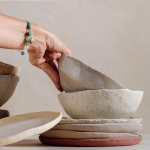 Può includere: Una collezione di ciotole e piatti in ceramica fatti a mano in varie dimensioni e tonalità neutre. Le ciotole hanno una texture maculata e sono impilate, con una mano che ne regola una. La tavolozza dei colori comprende beige, beige chiaro e bianco sporco.