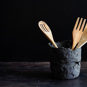 A black clay holder utensil with lots of texture contains three wooden ladles. It is made with strips of clay randomly joined together. The background is black and the table is made of wood. Dark Mood Photo.