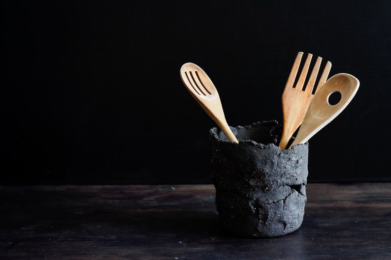 A black clay holder utensil with lots of texture contains three wooden ladles. It is made with strips of clay randomly joined together. The background is black and the table is made of wood. Dark Mood Photo.