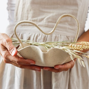 A woman in an apron holds a mottled beige stoneware basket in the shape of waves. It also has a wavy handle. It contains dowels.