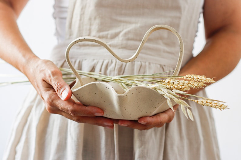 A woman in an apron holds a mottled beige stoneware basket in the shape of waves. It also has a wavy handle. It contains dowels.