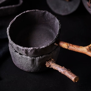 May include: Two black ceramic bowls with rough, textured exteriors. The bowls are stacked and have natural twig handles.