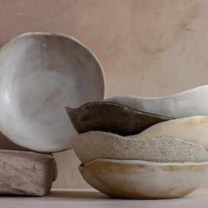 May include: A stack of handmade ceramic bowls in varying neutral tones. The bowls have organic shapes and textures, with a large, shallow bowl in the background. The color palette includes shades of white, beige, and brown, creating a rustic aesthetic.