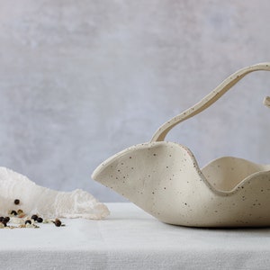 Front view of part of a basket on a white tablecloth. The basket is made of mottled beige stoneware. The handle is formed by two strips that meet in the center in the form of two spirals. A salt grinder can be seen on the left.