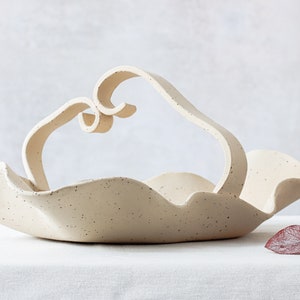 Front view of a basket on a white tablecloth. The basket is made of mottled beige stoneware. The handle is formed by two strips that are joined in the center in the form of two spirals.