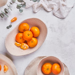 Still life photo. An oval-shaped fruit bowl of beige mottled clay is in the center, surrounded by pieces of the same material that look cut. The fruit bowl contains whole tangerines and tangerine segments. The other bowls also contain mandarins.