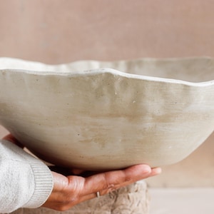 Könnte beinhalten: Eine große, cremefarbene Keramikschale mit gewelltem Rand. Die Schale hat eine strukturierte Oberfläche und wird von einer Hand gehalten. Die neutrale Farbe und die organische Form deuten auf eine rustikale oder minimalistische Ästhetik hin.