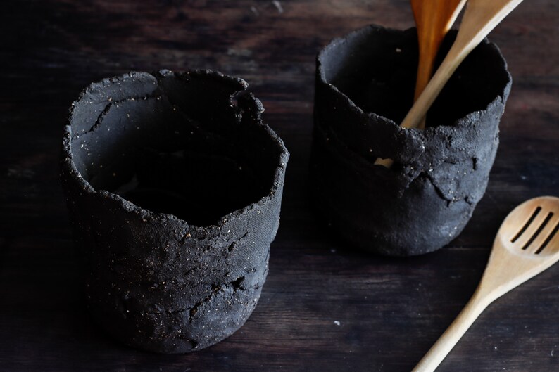 45º view of two dark utensil jars. They are made with textured black clay and hand-cut clay strips. It has holes. Three ladles are seen, two in one of the utensil holders and the other on the wooden table.