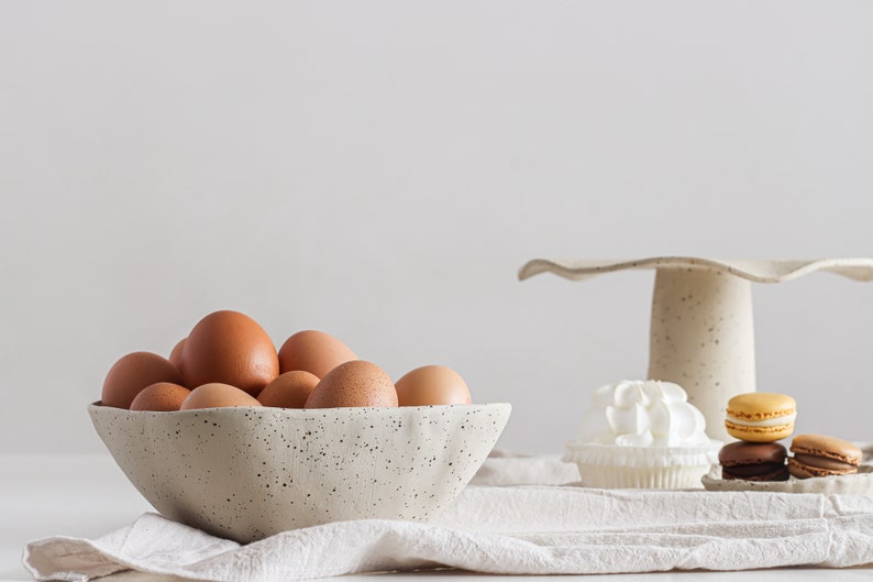 In the foreground a mottled beige stoneware bowl contains eggs. It sits on a beige tea towel. In the background is a cake stand and on the table are sweets and macarons.