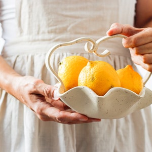 A woman in a beige apron is seen holding a wavy basket of mottled beige stoneware with a wavy handle. It contains three lemons.