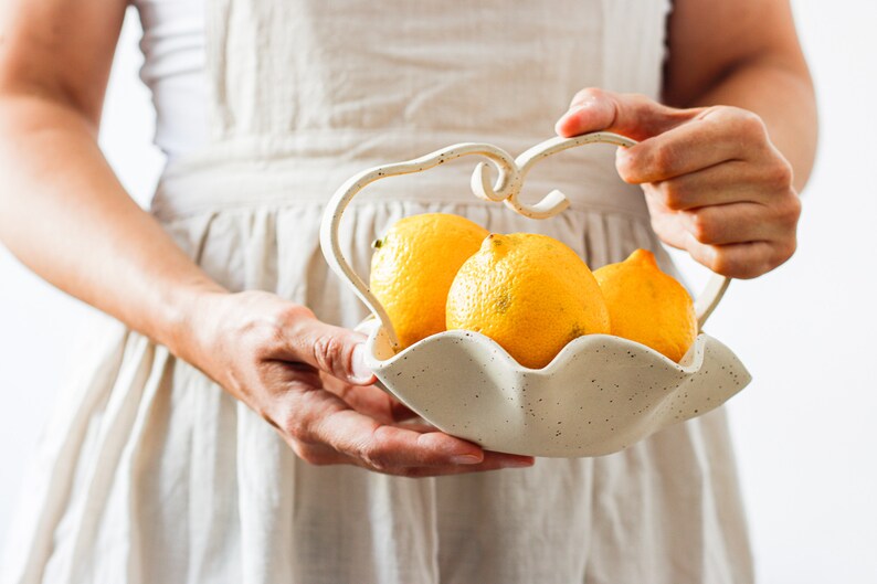 A woman in a beige apron is seen holding a wavy basket of mottled beige stoneware with a wavy handle. It contains three lemons.