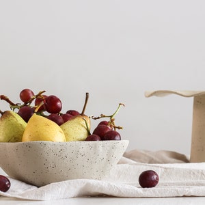 Still life scene in high-key. It shows a mottled beige stoneware bowl full of fruits. Behind is a cake stand of the same material. The base of the cake stand is cone-shaped. The part of the plate has many waves.