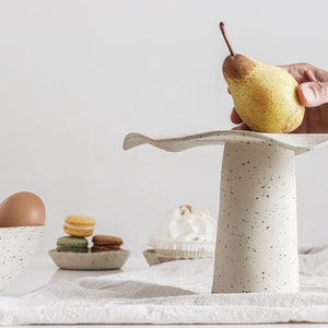 On the right is a cake stand with a hand holding a pear resting on it. On the left and in the background is a bowl with eggs, and two small plates with sweets. High-key photo.