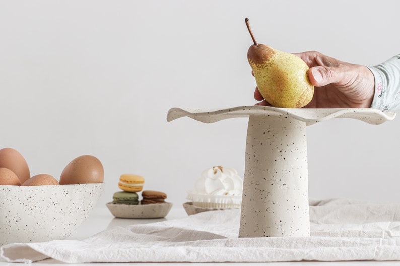 On the right is a cake stand with a hand holding a pear resting on it. On the left and in the background is a bowl with eggs, and two small plates with sweets. High-key photo.