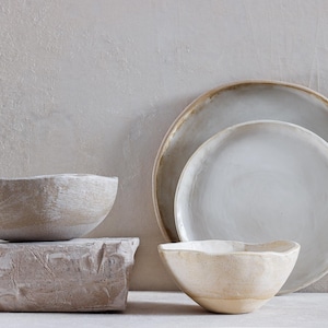 May include: A still-life composition featuring handmade ceramic tableware. The arrangement includes a large plate, a smaller plate, and two bowls in neutral tones. The pieces have a rustic, textured appearance, suggesting a natural aesthetic. The bowls are placed on a textured block.